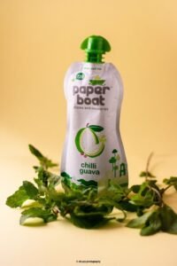 Paper Boat Chilli Guava drink pouch standing next to fresh green mint leaves on a warm yellow background.